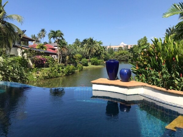A pool with a clear view of a river and surrounding greenery, including palm trees and a building in the background.