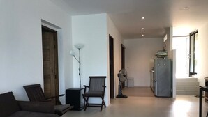 Ett modernt vardagsrum med en soffa, två fåtöljer, en golvlampa och en köksdel med ett kylskåp och en fläkt.