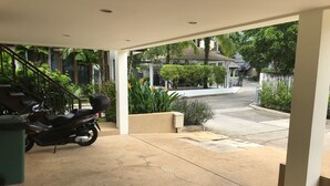 Ett takförsett utomhusområde med en motorcykel parkerad inuti, ett betonggolv och utsikt över en gata med hus och grönska.