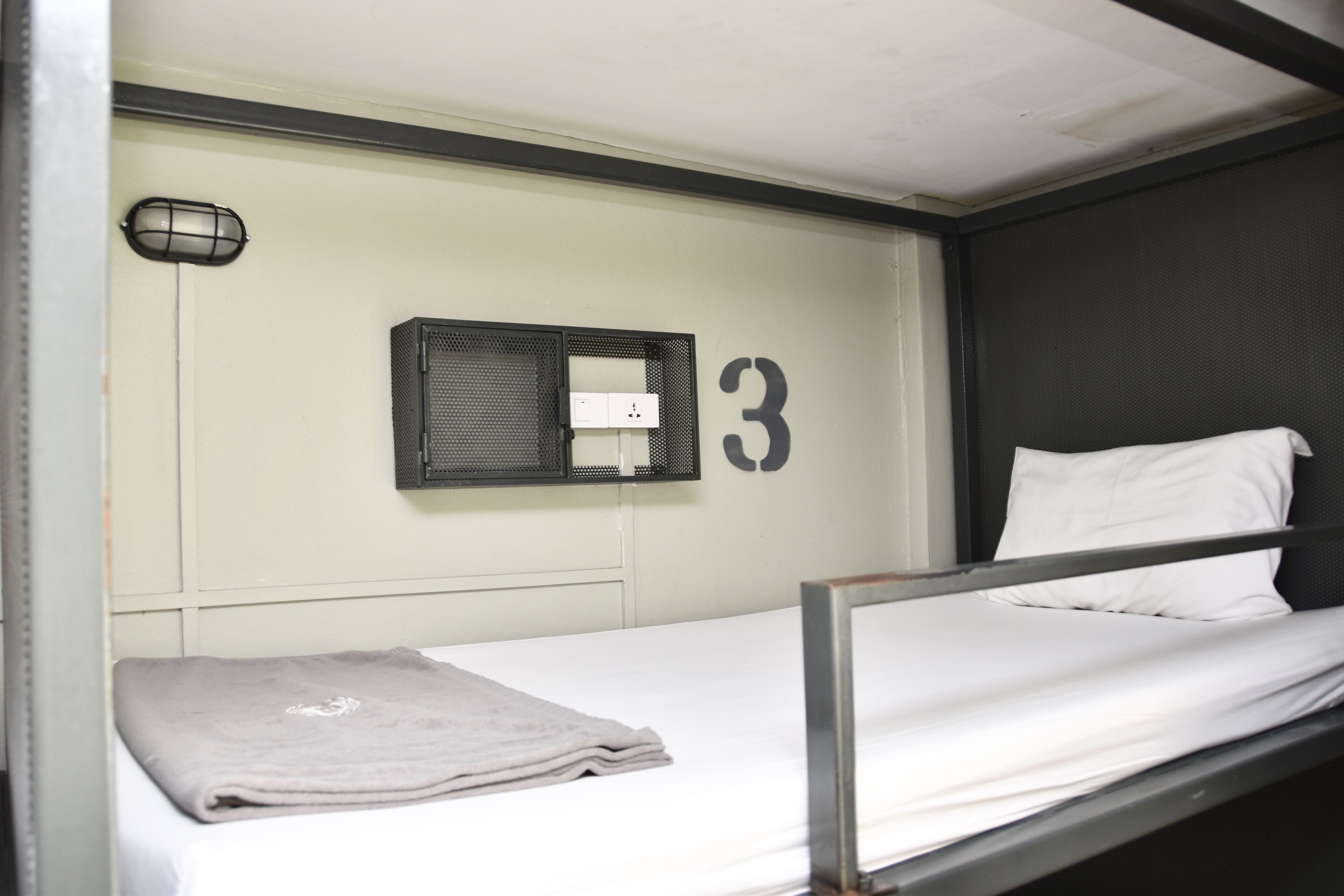 A bunk bed with a white mattress and a gray blanket. A small black-framed window is visible on the wall above the bed.