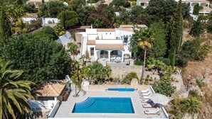 Una casa grande con piscina, rodeada de abundante vegetación y un muro de piedra.