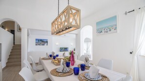 Un comedor con una mesa larga dispuesta para cuatro, sillas blancas, una lámpara de madera colgante y un cuadro con temática de playa en la pared.