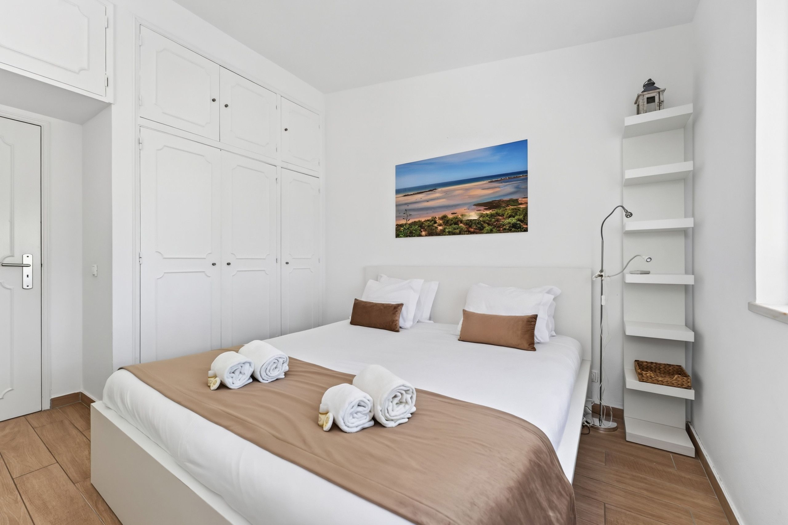 Quarto de hotel com cama, guarda-roupa branco, um quadro e mesa de cabeceira com abajur.