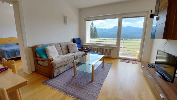 A living room with a sofa, a glass coffee table, a TV, and a view of mountains through large windows. - Holiday apartment in the resort Sonnenalm (Sonnenalm)