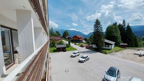 A parking lot with several cars, a building with a balcony, and a view of houses and mountains in the background. - Holiday apartment in the resort Sonnenalm (Sonnenalm)