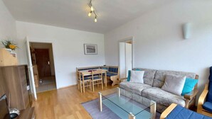 A modern living room with a glass coffee table, a sofa with cushions, a dining table with benches, and a kitchen area visible through an open door. - Holiday apartment in the resort Sonnenalm (Sonnenalm)