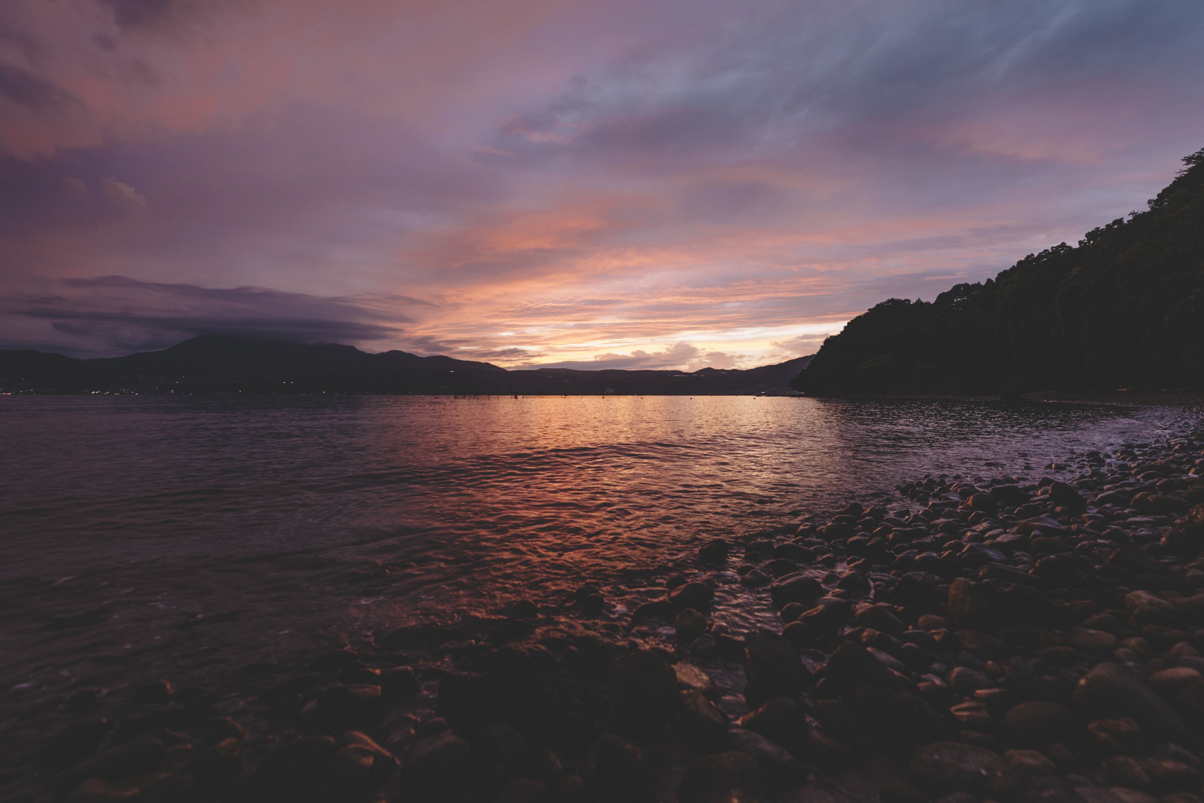 A calm lake at sunset with a rocky shoreline and distant mountains.