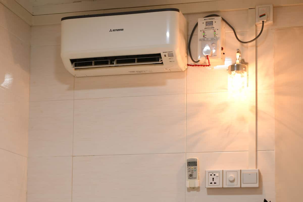 Une petite pièce bien éclairée, avec un lit, un climatiseur, un luminaire fixé au mur, une petite statue et une étagère avec une plante.