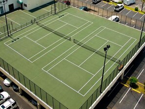 A tennis court with a surrounding fence, adjacent parking, and a building with a green roof. - Sands Beach Club II 327, Lovely 2 BR Ocean Front Condo with Indoor Outdoor Swimming Pool, Hot Tub and Kiddie Pool (MYRTLE BEACH)