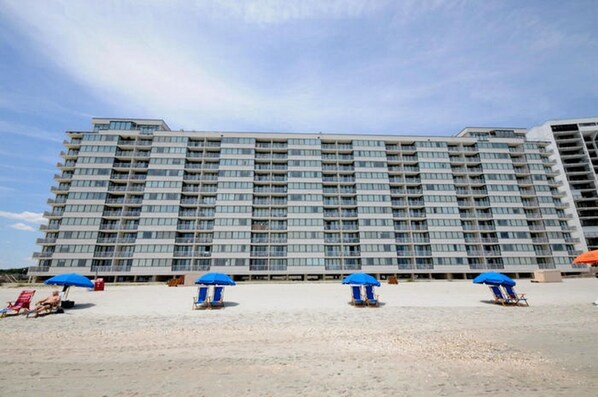 A high-rise hotel with multiple balconies and a sandy beach area with blue umbrellas. - Sands Beach Club II 327, Lovely 2 BR Ocean Front Condo with Indoor Outdoor Swimming Pool, Hot Tub and Kiddie Pool (MYRTLE BEACH)