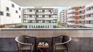 En balkon med to fletstole og et lille bord dækket til to, med udsigt over en moderne lejlighedsbygning med flere balkoner og vinduer.