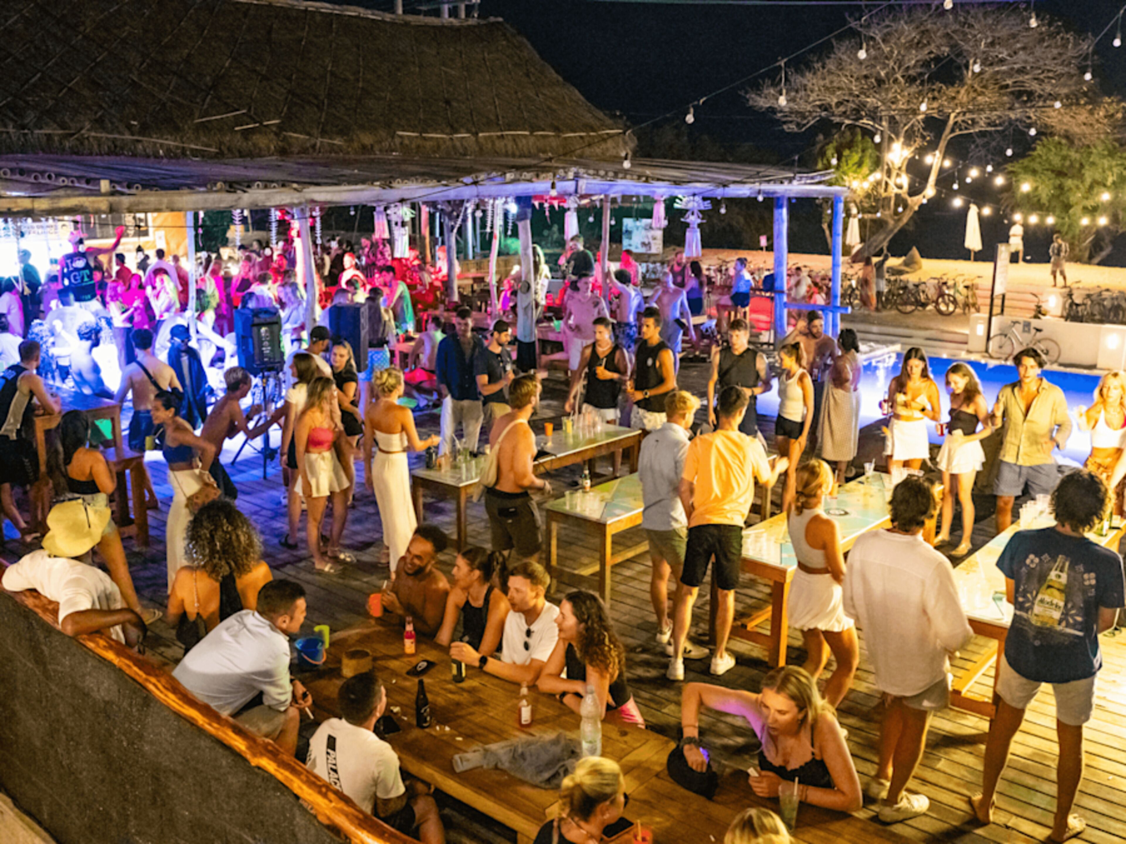 A crowded outdoor bar area with wooden tables and chairs, surrounded by a thatched-roof structure.