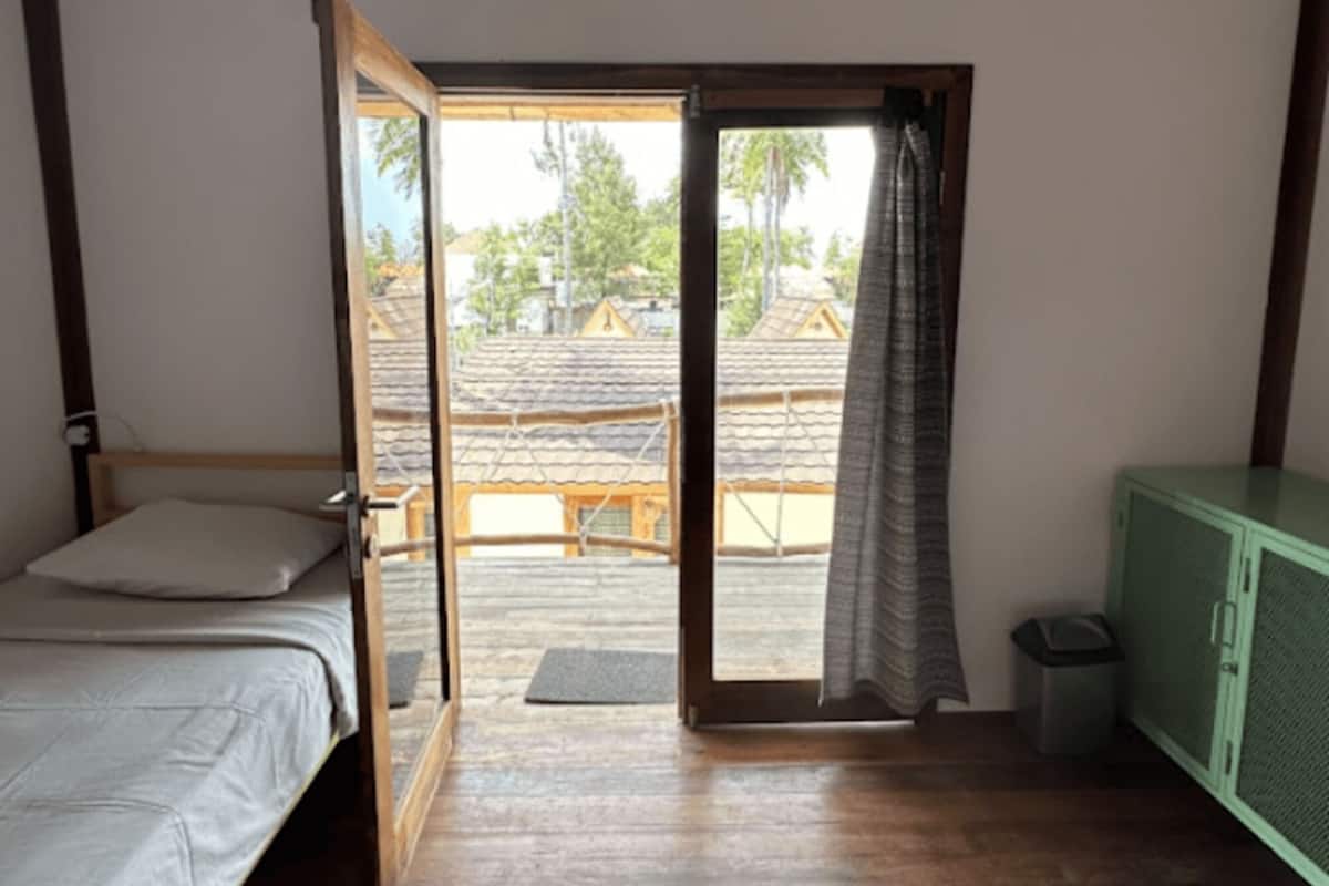 Ein Zimmer mit einem Bett, einem kleinen Schrank und einer Schiebetür zu einem Außenbereich mit Blick auf Häuser und Bäume.