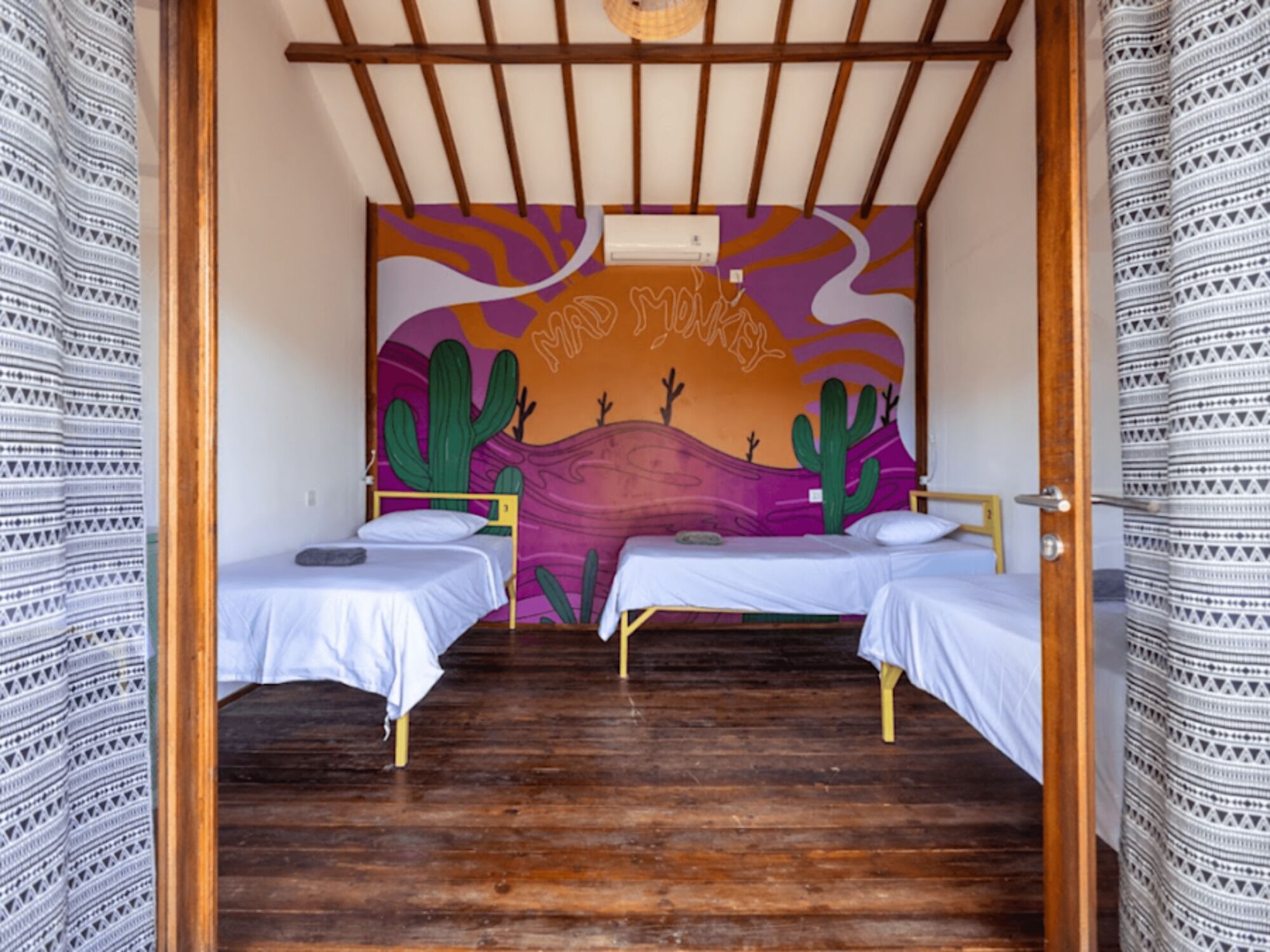A room with two single beds, wooden flooring, a mural with cacti, and a ceiling fan.
