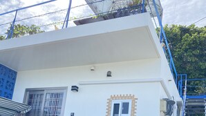 Un edificio de varios niveles con fachada blanca, un balcón con plantas y barandilla azul.