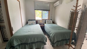 A room with two single beds, a window with blinds, an air conditioning unit, and a metal rack. - Sesoko Terrace 2 (Motobu)