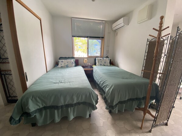 A room with two single beds, a window with blinds, an air conditioning unit, and a metal rack. - Sesoko Terrace 2 (Motobu)