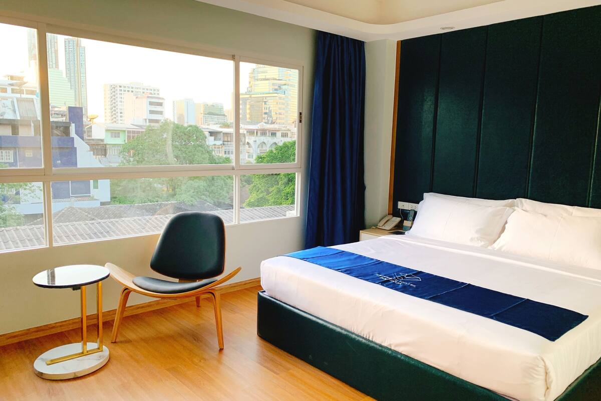 Une chambre d’hôtel avec un grand lit, une chaise, une petite table et une vue sur la ville grâce à de grandes fenêtres.