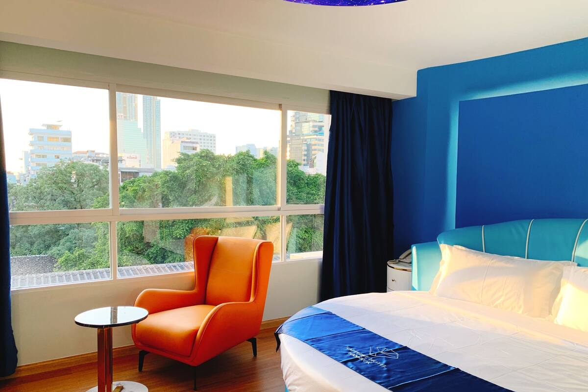 Une chambre d’hôtel avec un grand lit, une petite table ronde, une chaise orange et une vue sur les immeubles par la fenêtre.