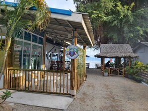 A beachside restaurant with a covered outdoor seating area, a reception desk, and a thatched-roof hut.