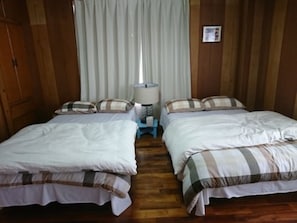 A room with two beds, wooden walls, a nightstand with a lamp, and a window with curtains. - Kokusaburo House (Onna)