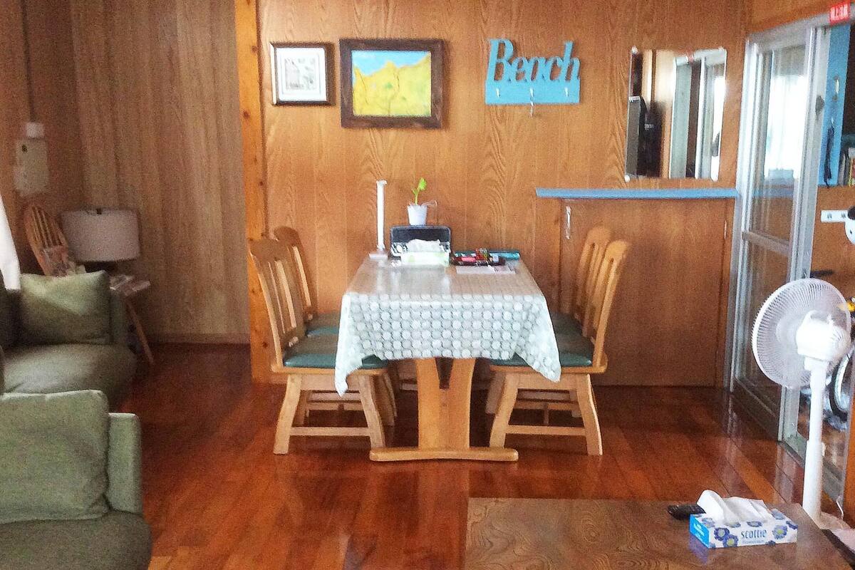 Un comedor de madera con una mesa y sillas, una mesita con lámpara, un ventilador y un letrero que dice 