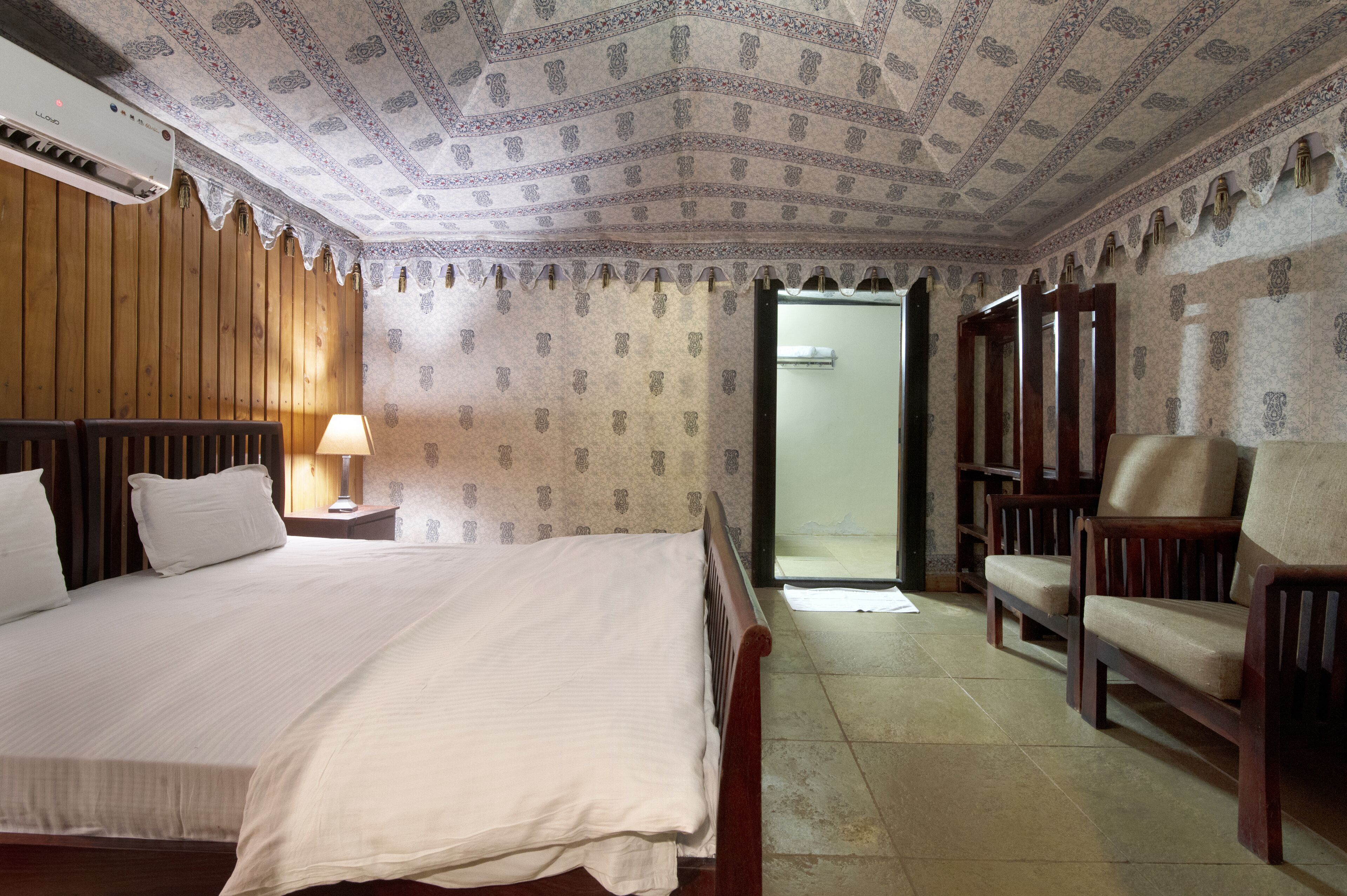 A hotel room with a large bed, a wooden headboard, a nightstand with a lamp, a wooden chair, and a wardrobe.