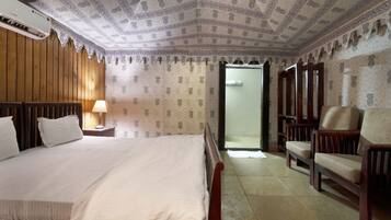 A hotel room with a large bed, a wooden headboard, a nightstand with a lamp, a wooden chair, and a wardrobe.