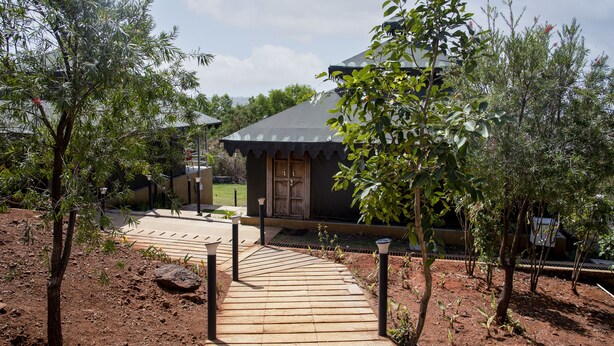 Um edifício com telhado escuro e porta de madeira, rodeado por árvores e um caminho pavimentado.