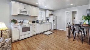 A modern kitchen with white cabinets, a microwave, stove, and a refrigerator. There is a dining area with a wooden table and chairs. - The West End Cottage (Buhl)