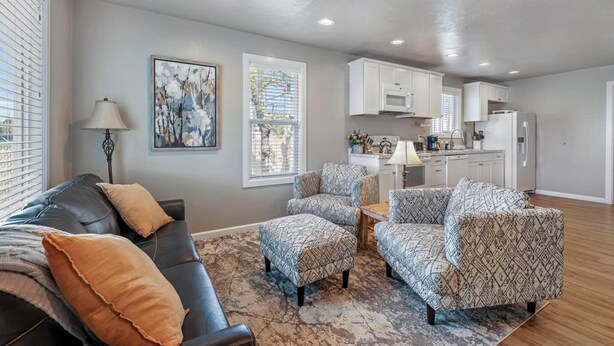 A living room with a black leather sofa, patterned armchairs, and a coffee table. There is a kitchen in the background with white cabinets and a refrigerator. - The West End Cottage (Buhl)