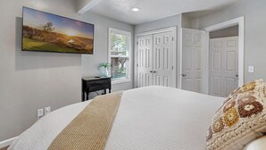 A bedroom with a large bed, a flat-screen TV mounted on the wall, a window with blinds, and a door. - The West End Cottage (Buhl)