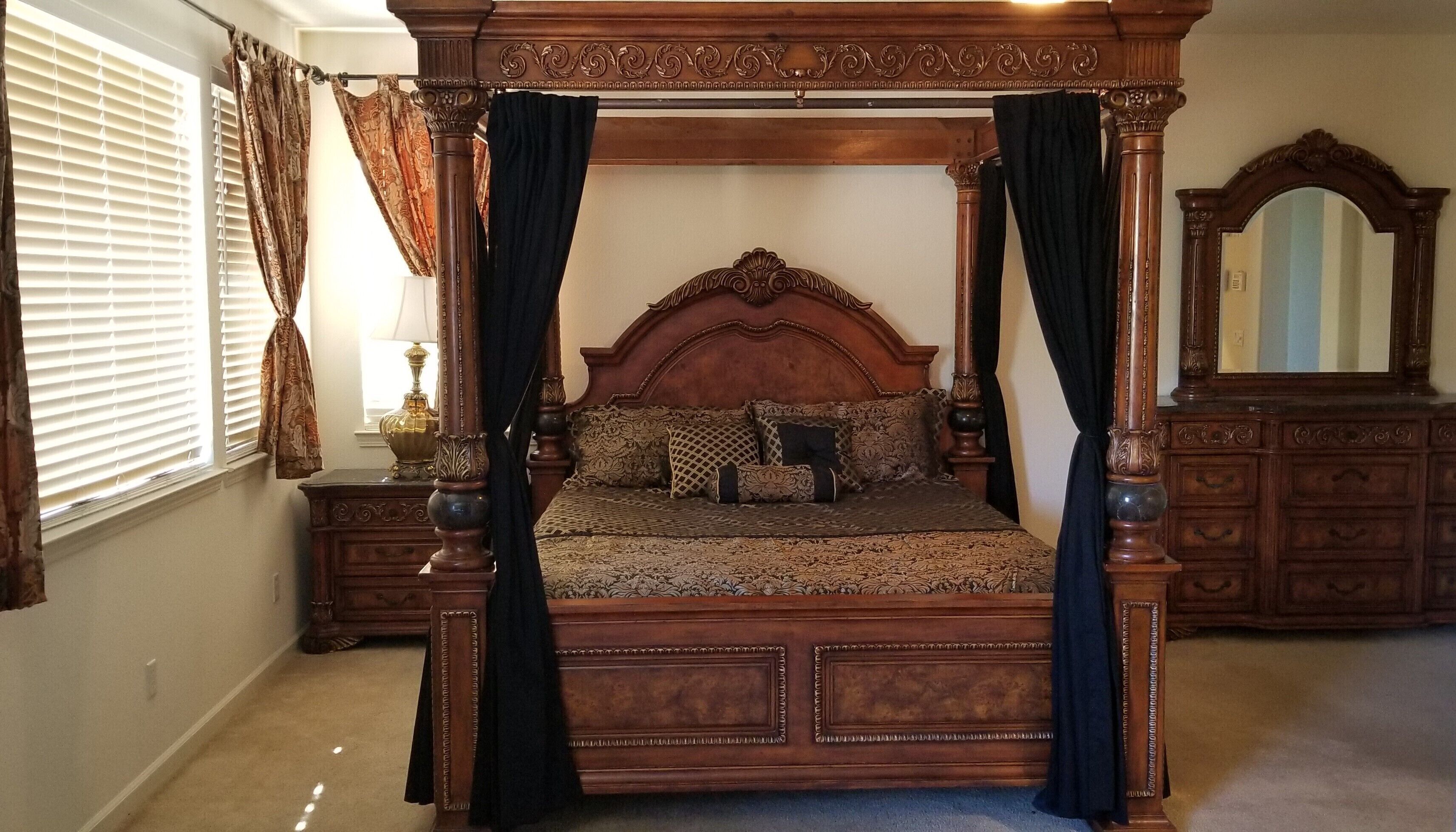 Una cama con dosel y pilares, con cabecera de madera y cortinas oscuras, flanqueada por mesitas de noche con lámparas y un aparador con espejo.