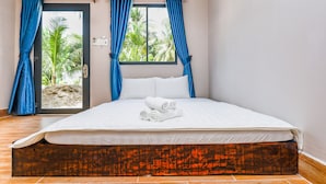 Una cama bien hecha con sábanas blancas y toallas enrolladas, un cabecero de madera y vistas a palmeras a través de una ventana con cortinas azules.