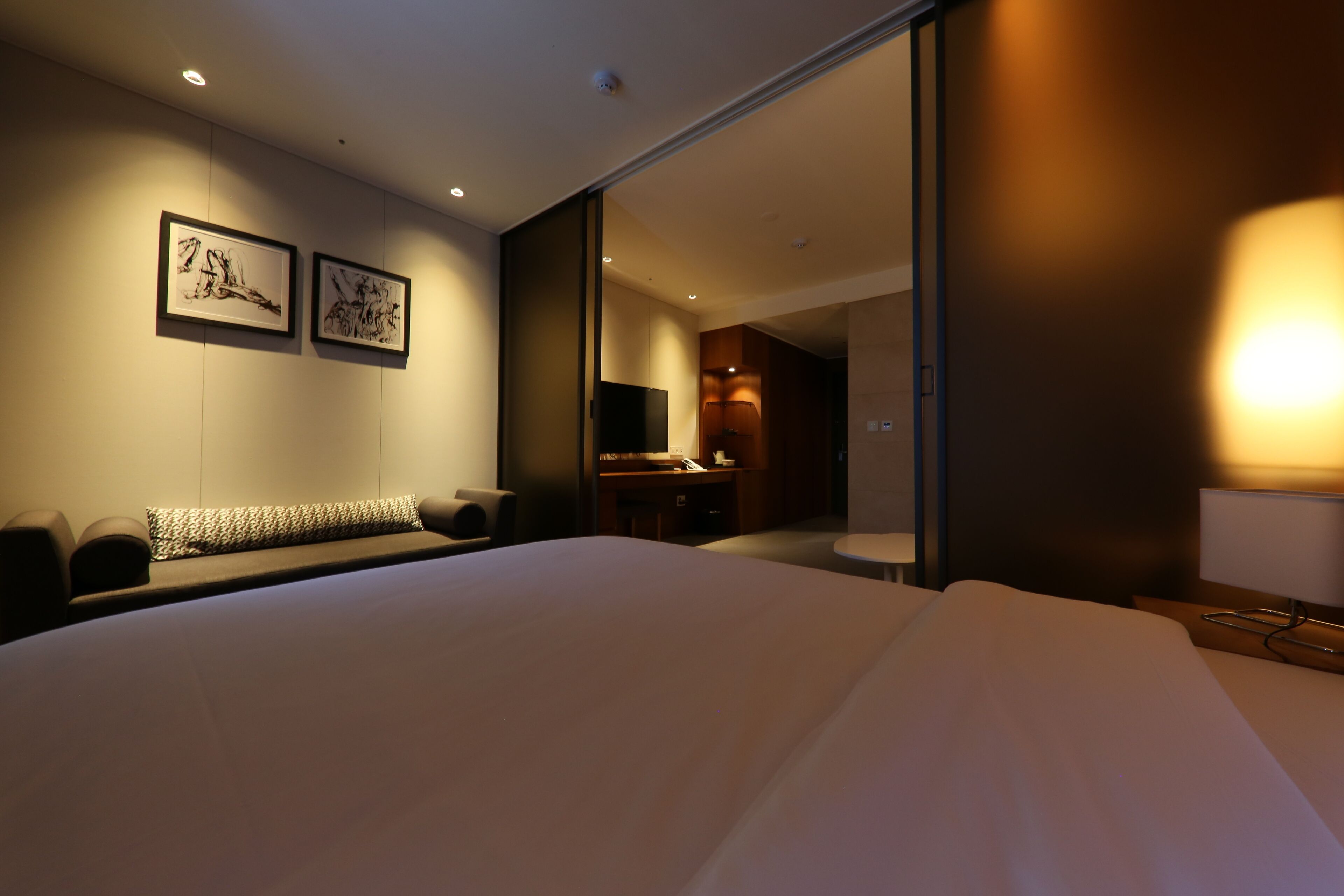 Una habitación de hotel moderna con una cama grande, una zona de estar con sofá y cojines, un escritorio con lámpara, y dos obras de arte enmarcadas en la pared.