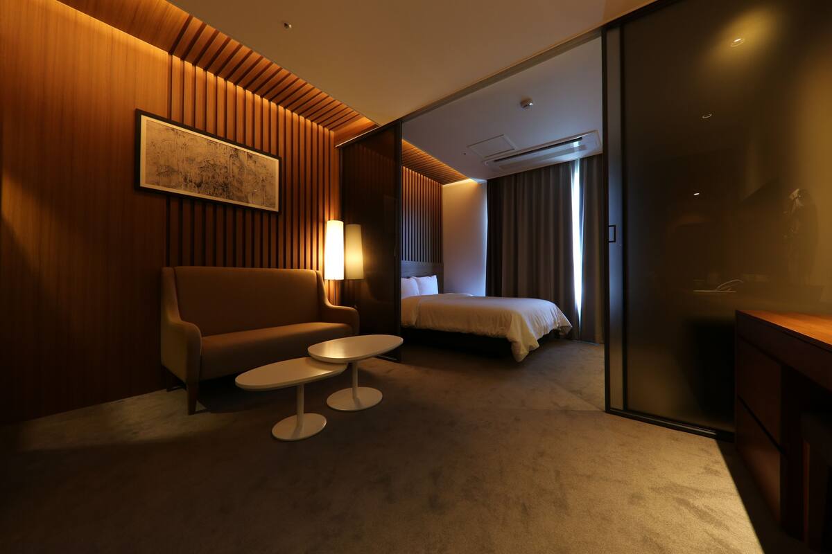 Una habitación de hotel con sofá, dos mesitas, cama y un amplio ventanal.