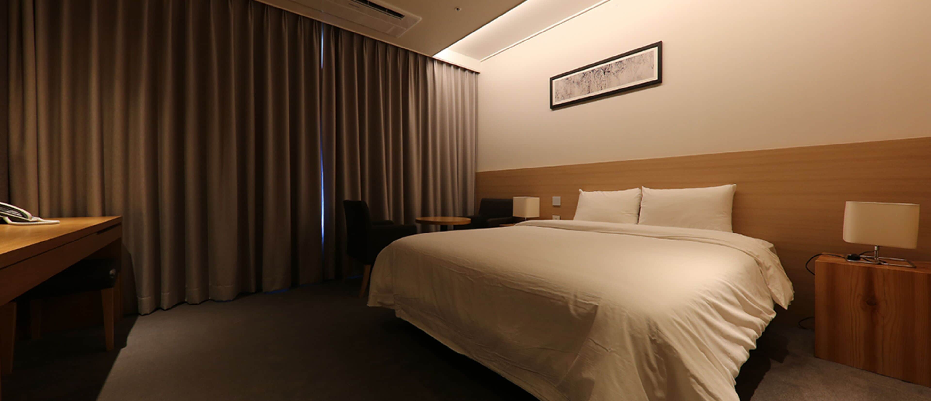 A hotel room with a large bed, a desk, a chair, a bedside table with a lamp, and a framed picture on the wall.