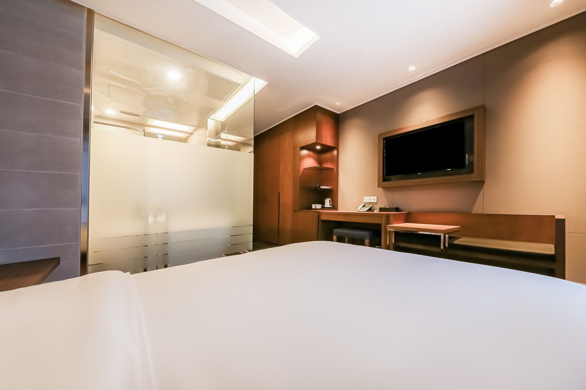 Una habitación de hotel moderna con una cama grande, un escritorio con silla, un televisor de pantalla plana montado en la pared y una mampara de vidrio.