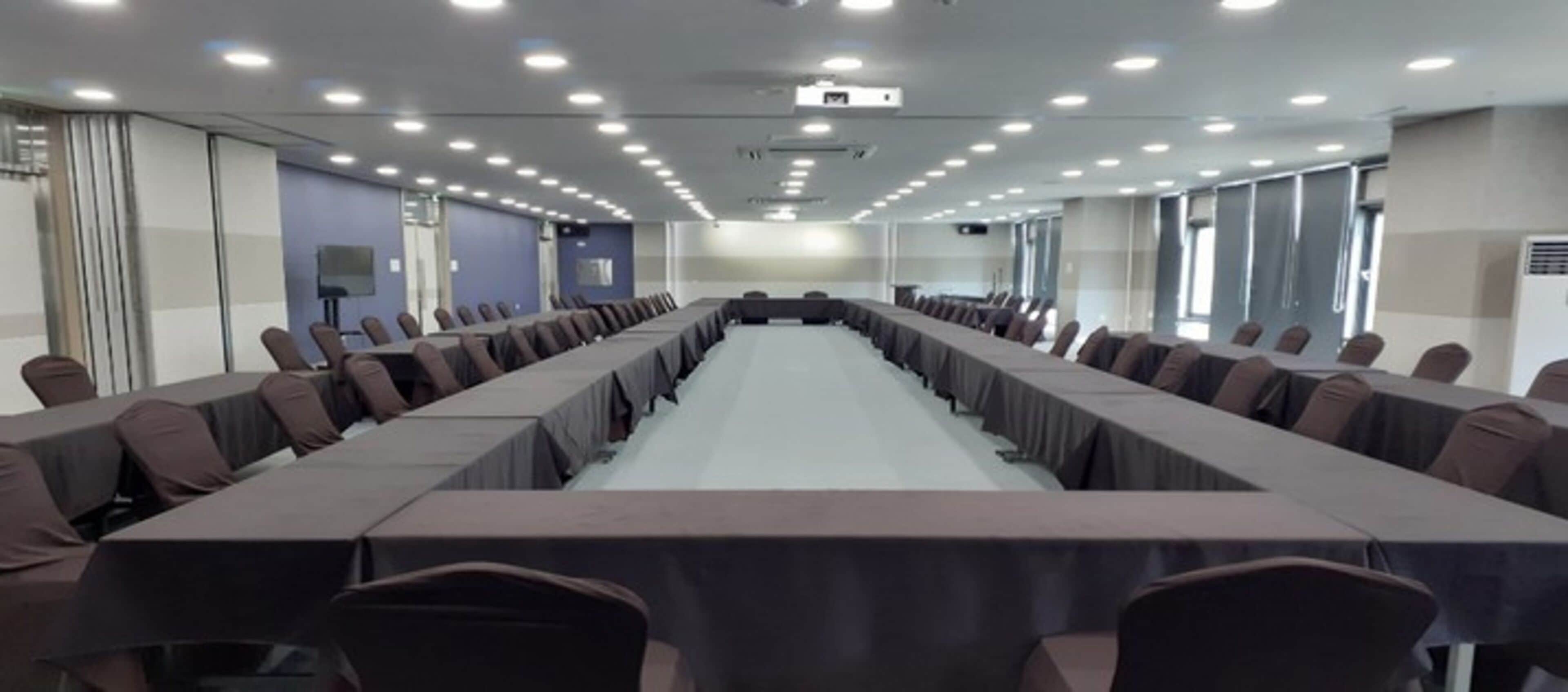 A conference room with a long table and chairs, equipped with a projector and ceiling lights.
