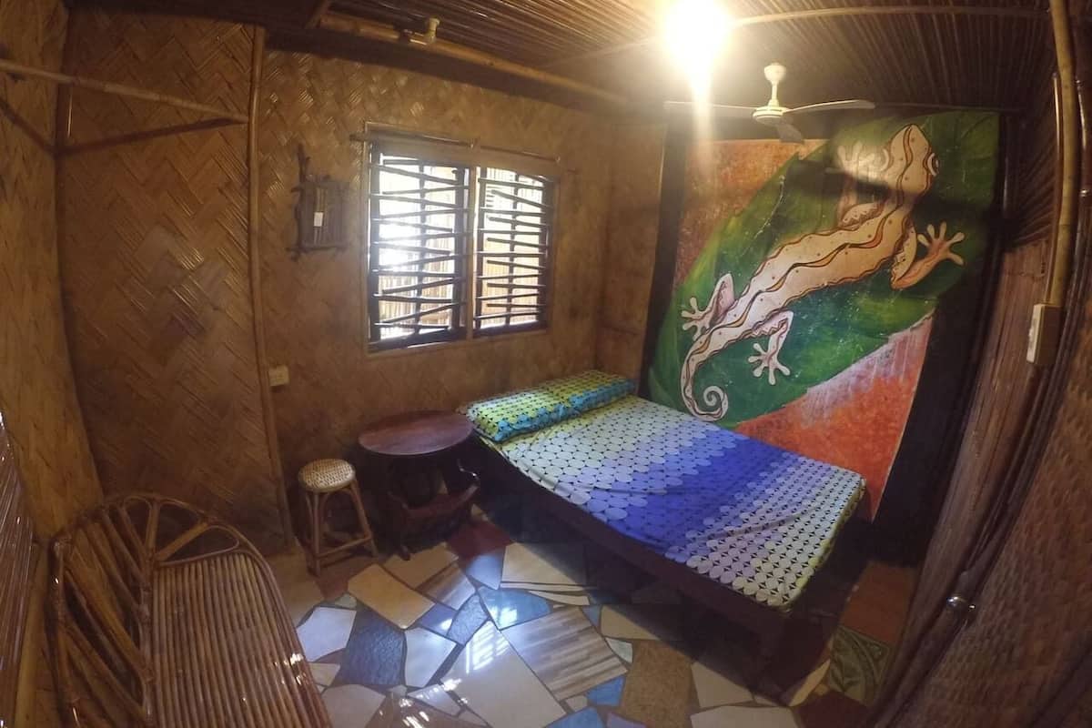 Ein Zimmer mit einem Bett, einem Hocker, einem Fenster und Wandbild.