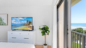 A bedroom with a TV on a white dresser, a potted plant, and a balcony with a view of the beach and ocean. - Bright & Modern Penthouse w/ Stunning Ocean Views, Heated Pool & BBQ Grills! (Cocoa Beach)