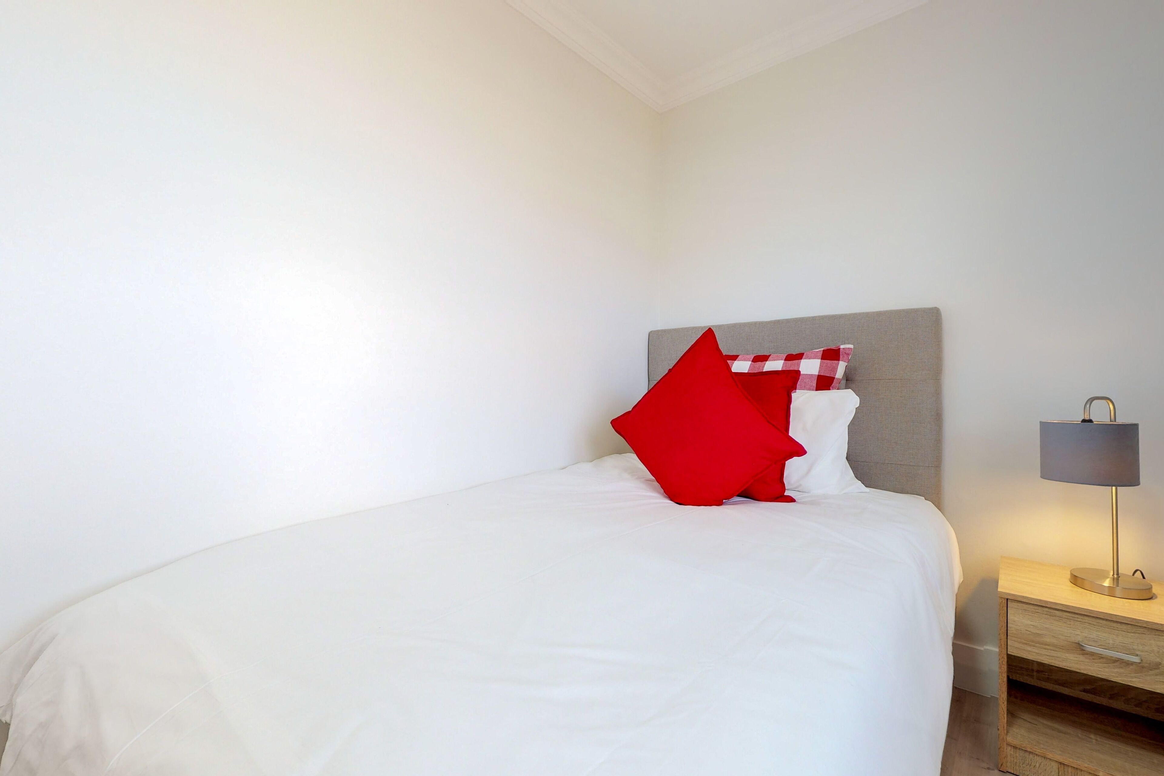 Un lit bien fait, avec une literie blanche et un oreiller rouge, une table de chevet avec une lampe et une commode en bois.