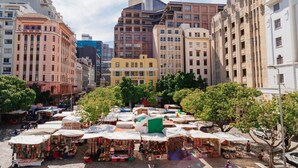 Um mercado ao ar livre movimentado, com vários quiosques sob guarda-sóis, cercado por edifícios históricos.