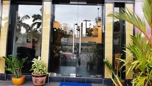 The entrance of Hotel Cappuccino with a 'Welcome' mat, potted plants, and a clear glass facade.