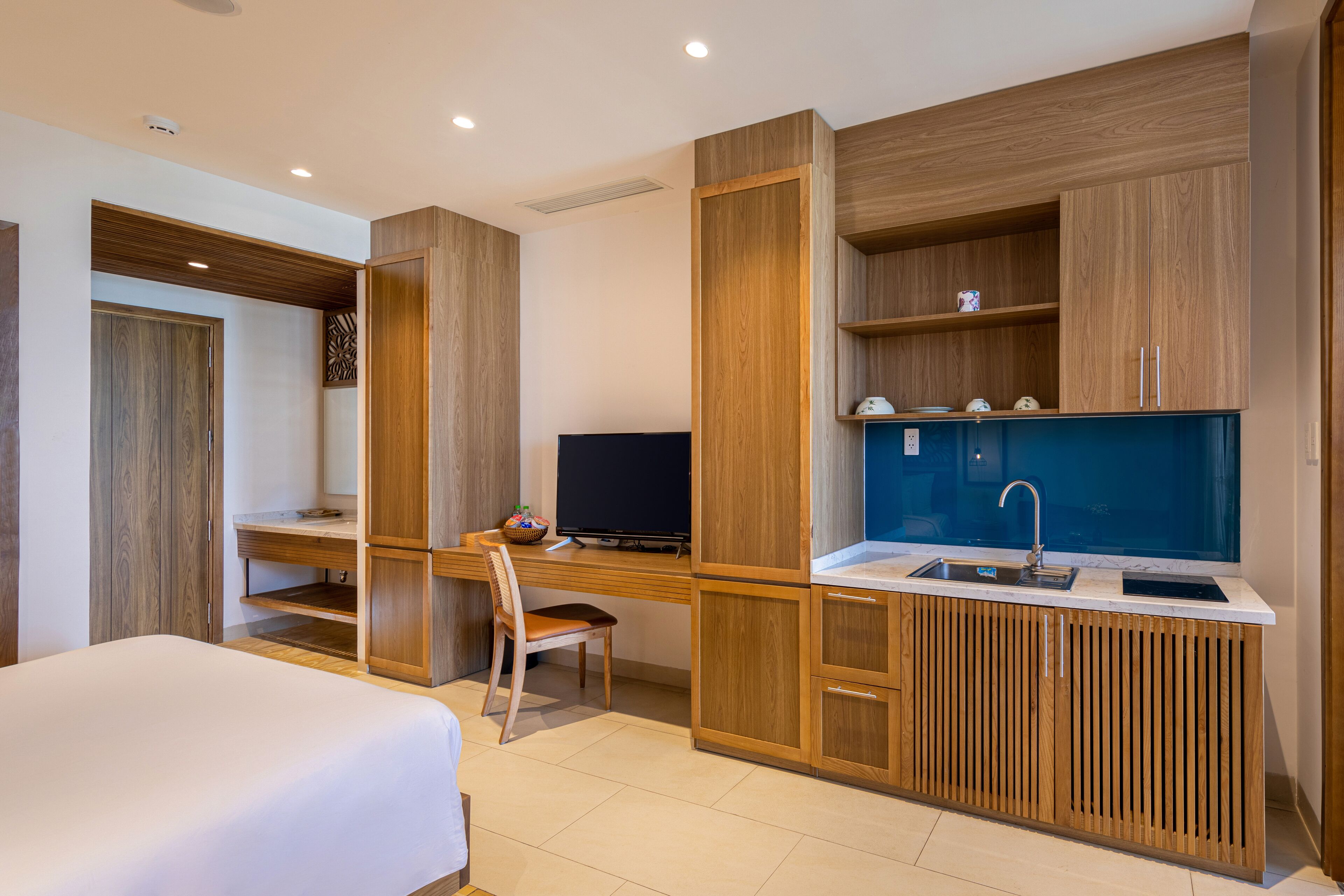 Une chambre d’hôtel moderne équipée d’un lit, d’un bureau avec un ordinateur, d’une chaise et d’une kitchenette comprenant un évier et des placards.