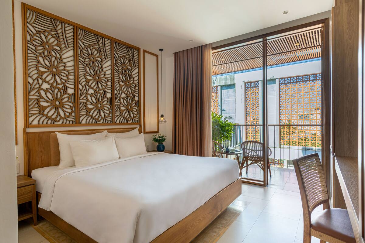Habitación de hotel con una cama grande, cabecera de madera, panel decorativo en la pared, balcón con vistas y zona de estar con sillas.