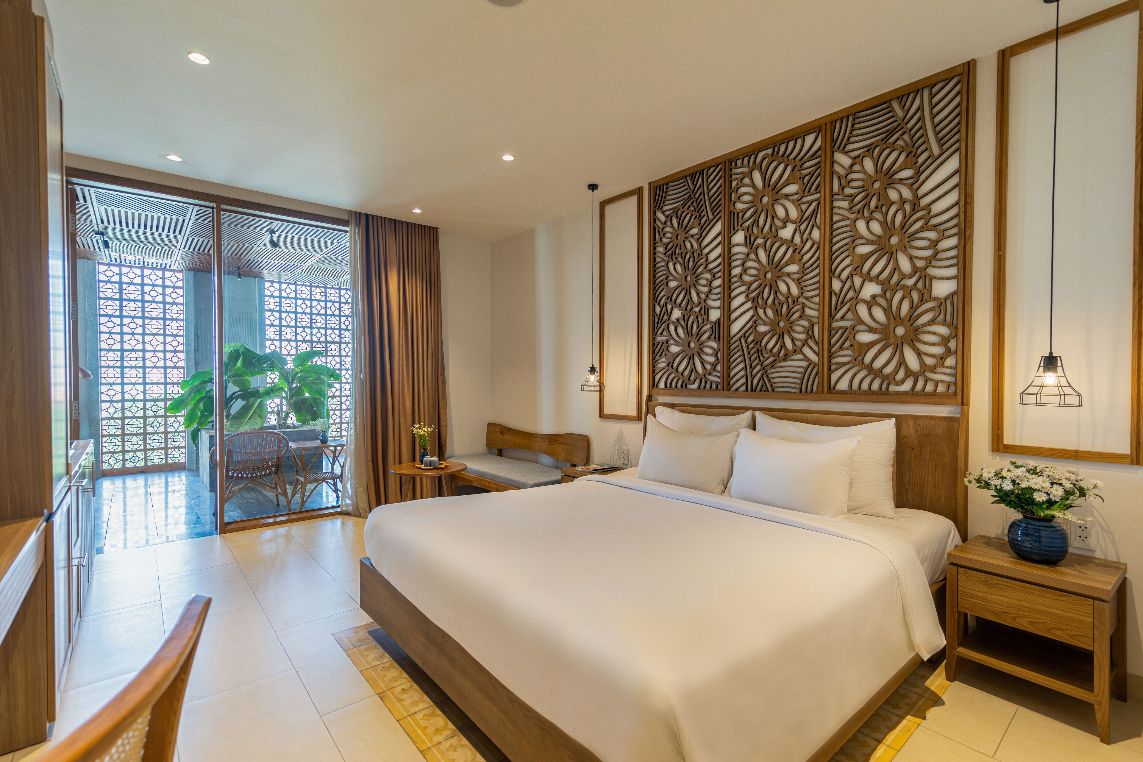 Une chambre d’hôtel avec un grand lit, une tête de lit en bois, un panneau décoratif au mur, une petite table avec un vase, et une vue sur un espace extérieur verdoyant, accessible par une porte vitrée.