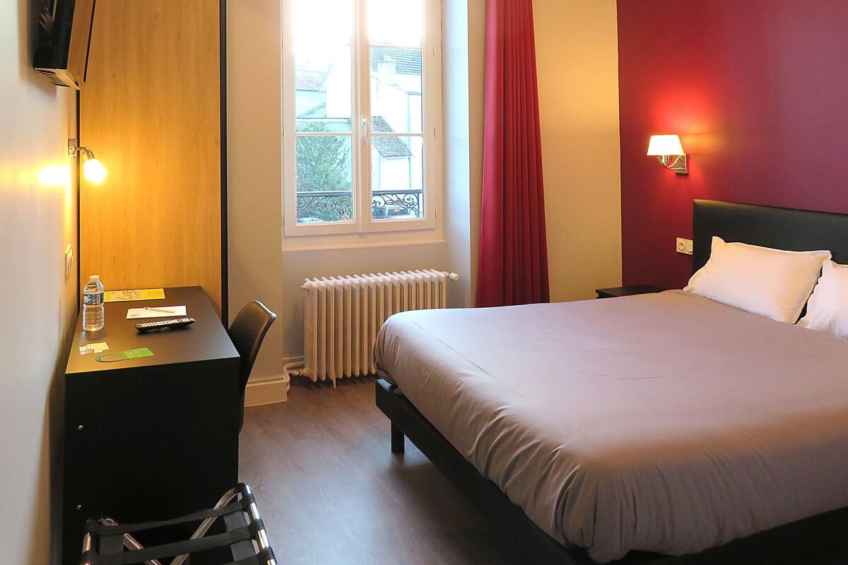 A hotel room with a bed, a desk with a chair, a television mounted on the wall, a window with curtains, and a radiator.