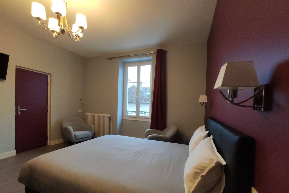 A hotel room with a bed, a chair, a window with curtains, a radiator, and a wall-mounted lamp.