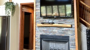 Un salon avec une cheminée en pierre, un téléviseur fixé au mur et une horloge accrochée au mur.